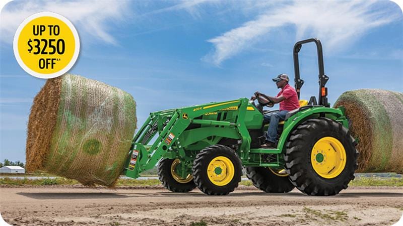 Man in the seat of a John Deere 4044M tractor with bay spear moving bale of hay.