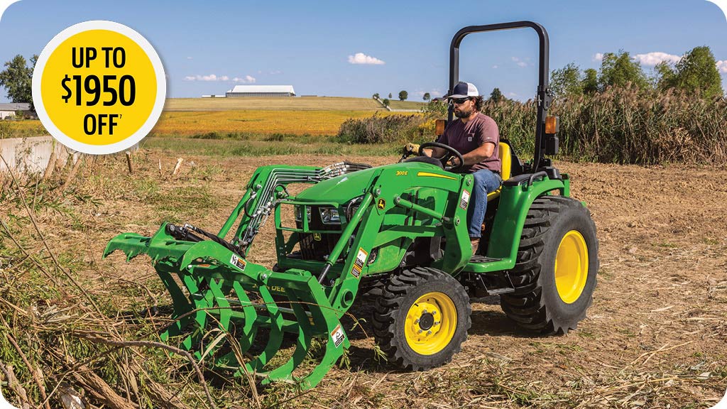 Man in the seat of a John Deere 3E tractor with grapple moving brush.