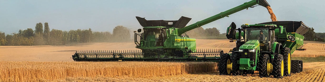 Une moissonneuse-batteuse verte récolte du blé et décharge le grain à la volée dans une remorque tirée par un tracteur, au milieu d’un champ doré.