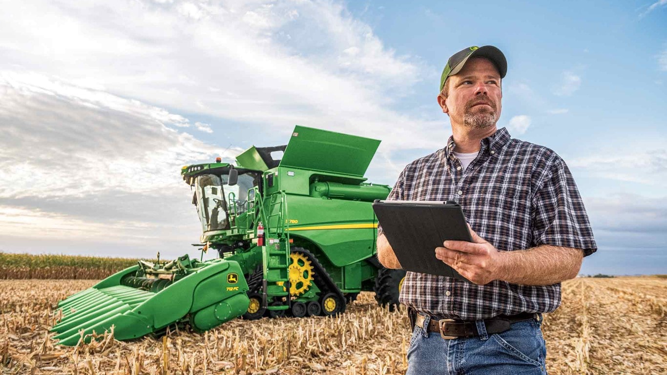 Un agriculteur vêtu d’une chemise à carreaux et d’une casquette, tablette à la main, devant une moissonneuse-batteuse verte dans un champ.
