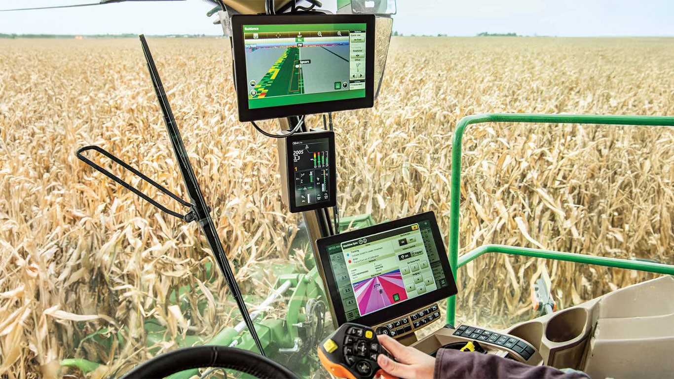 Vue dans la cabine des consoles John Deere installées dans une moissonneuse-batteuse, avec la main de l’utilisateur sur le levier de commande et du maïs à maturité visible à travers la vitre.