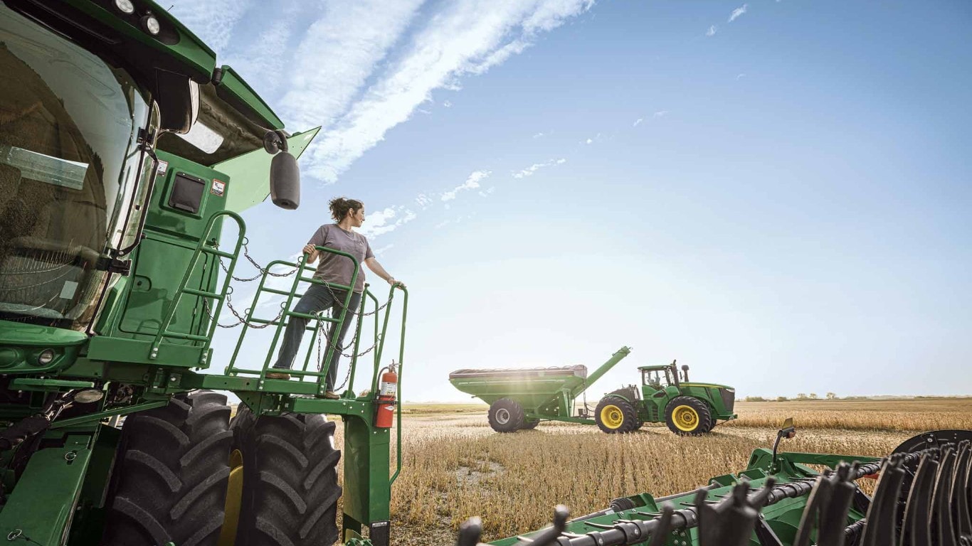 Sur la plate-forme arrière d’une moissonneuse-batteuse verte, une utilisatrice observe un chariot à grain et un tracteur dans un champ de blé, sous un ciel bleu. Sur la plate-forme arrière d’une moissonneuse-batteuse verte, une utilisatrice observe un chariot à grain et un tracteur dans un champ de blé, sous un ciel bleu.