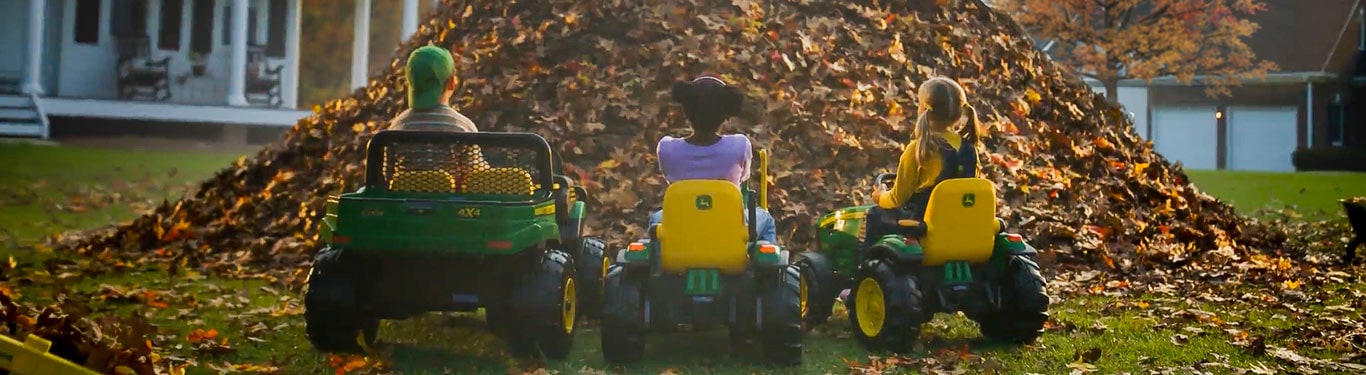 Kids sitting on John Deere machines in front of a leaf pile on a crisp autumn day