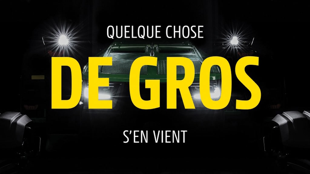 Image publicitaire noire montrant une machine qui sort de l'ombre phares allumés et affichant le texte « QUELQUE CHOSE DE GROS S'EN VIENT ».