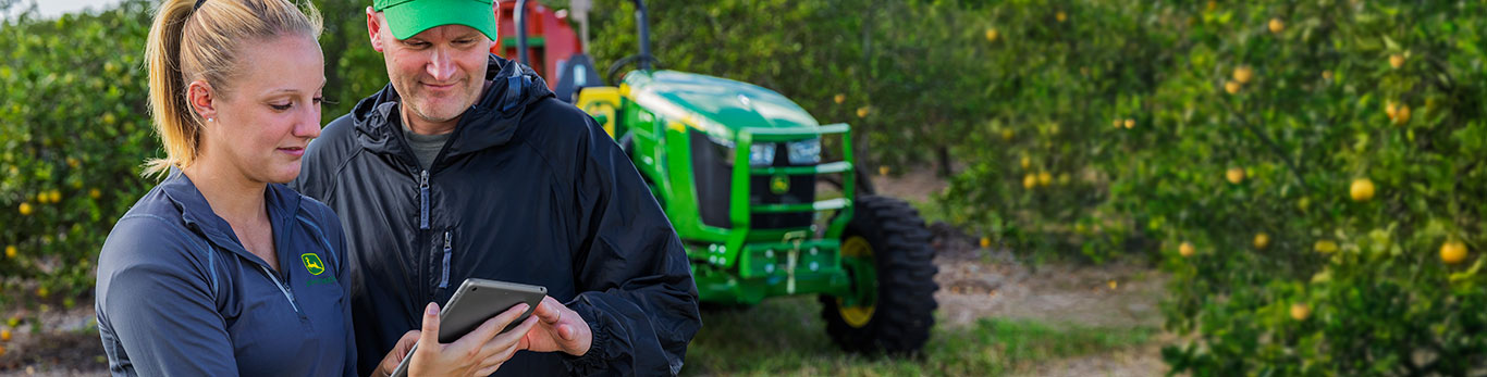Une femme et un homme regardant une tablette devant un tracteur John Deere dans un verger.