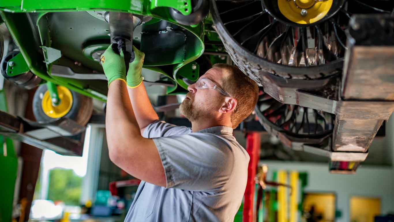 Un mécanicien portant des gants et des lunettes de protection John Deere travaille sous une tondeuse John Deere