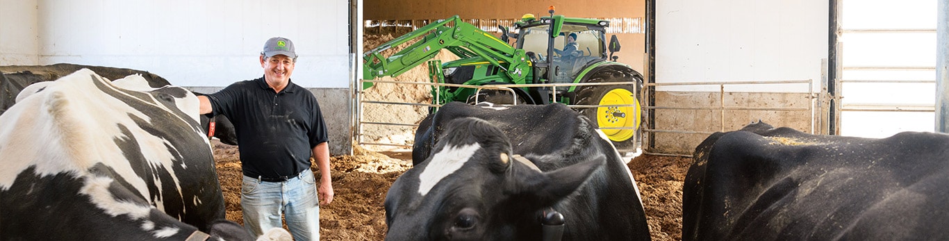Agriculteur dans la grange avec des vaches noires et blanches ; tracteur vert avec des roues jaunes en arrière-plan.