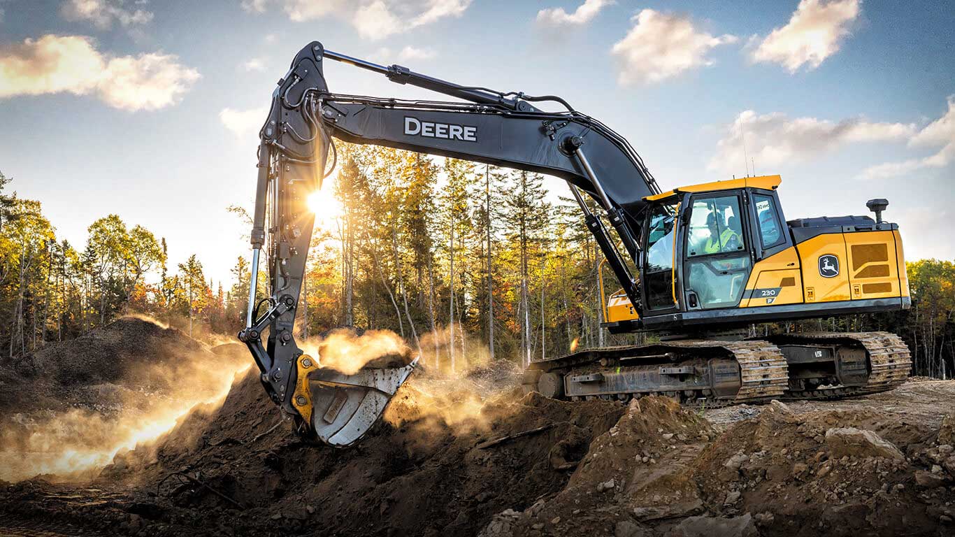Excavatrice de John&nbsp;Deere creusant le sol sur un chantier de construction, la lumière du soleil filtrant à travers les arbres en arrière-plan.
