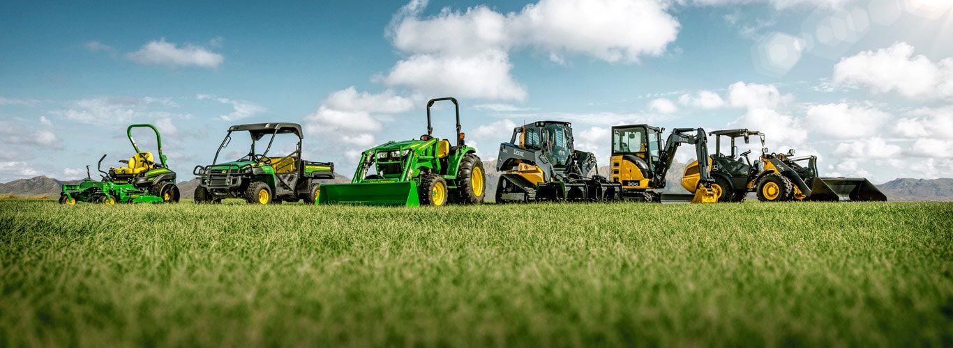 Gamme d'équipement pour travaux paysagers et d'entretien des terrains de John Deere dans un champ d'herbe avec des montages au loin