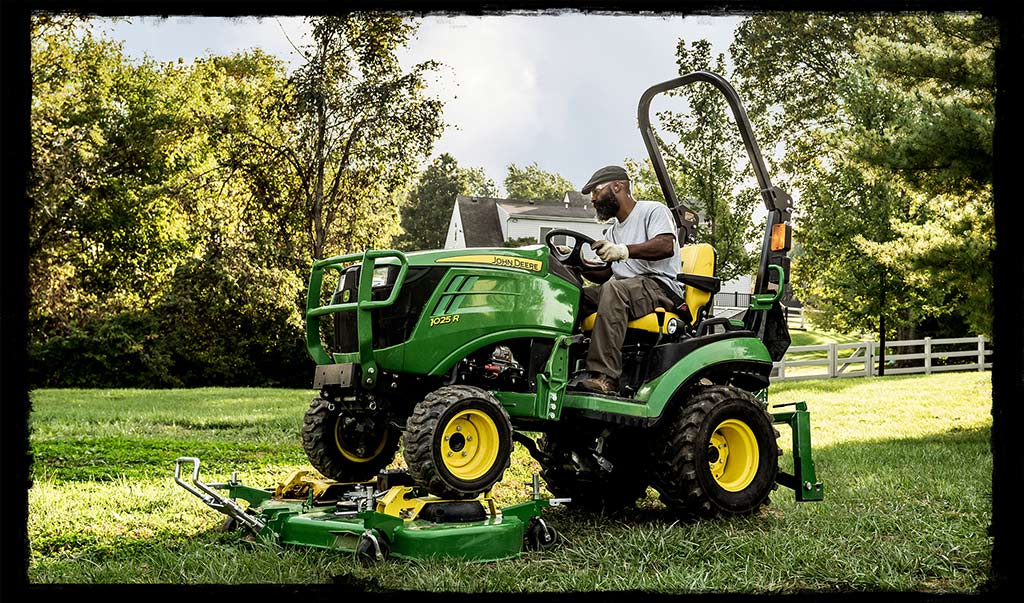 Un homme sur un tracteur jaune et vert de la série 1 de John Deere se trouve dans une cour à la pelouse verte avec une clôture et de grands arbres en arrière-plan.