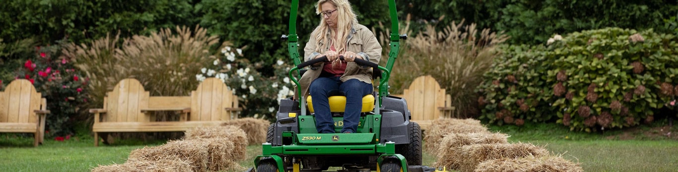 Une femme aux cheveux blonds vêtue d’une chemise grise et d’un jeans conduit une tondeuse verte et jaune John&nbsp;Deere coupant l’herbe. Un banc en bois est installé en arrière-plan et des balles de foin sont installées de chaque côté de la tondeuse.