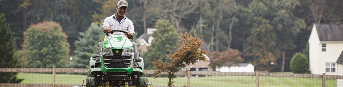 Un homme à barbe grise vêtu d’une chemise gris clair et d’une casquette de baseball noire se déplace sur une tondeuse verte et jaune de John Deere sur un terrain à pelouse avec une clôture en bois en arrière-plan.