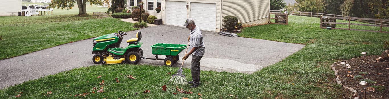 A man in a gray collared shirt and pants with a gray and white baseball cap on rakes his green grass yard. There is a green and yellow lawn mower with a wheelbarrow attached to the back in the background.