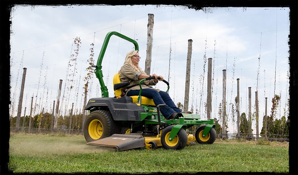 Une femme aux cheveux blonds vêtue d'une chemise grise et d'un jeans bleu conduit une tondeuse verte et jaune John Deere coupant l’herbe.