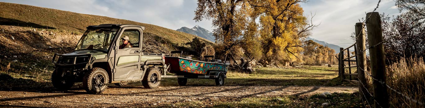 Woman in a dark green and black John Deere Gator pulls a bright multicolored boat with a mountain range in the background.