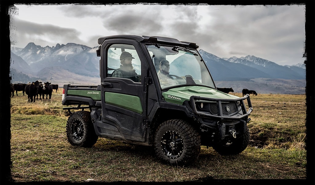 Un véhicule utilitaire John Deere noir et vert foncé roule dans l’herbe, les silhouettes de deux personnes étant visibles à l’intérieur de la cabine. Les montagnes enneigées et les vaches sont en arrière-plan.