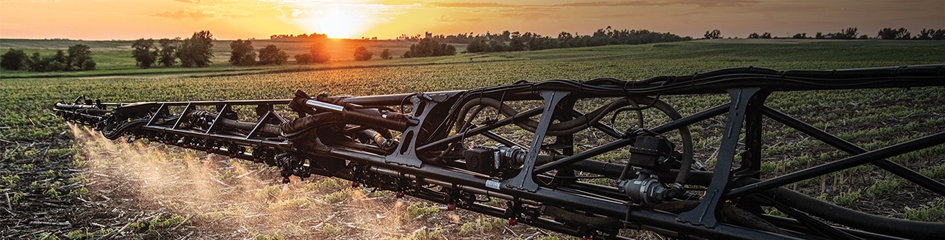 A sprayer boom applying a fine mist across a field at sunset.