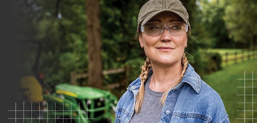 Une personne portant une veste en denim et une casquette, debout à l’extérieur, avec un tracteur compact John Deere en arrière-plan.