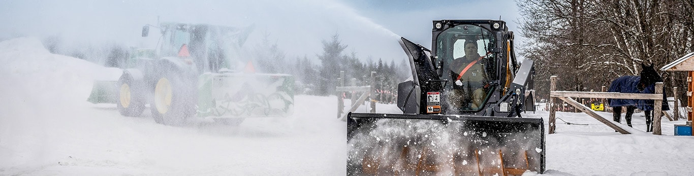 Personne soufflant de la neige à l’aide d’une chargeuse à direction différentielle et d’une souffleuse à neige avec un tracteur utilitaire de la série 5 équipé d’une souffleuse à neige et d’une chargeuse en arrière-plan.