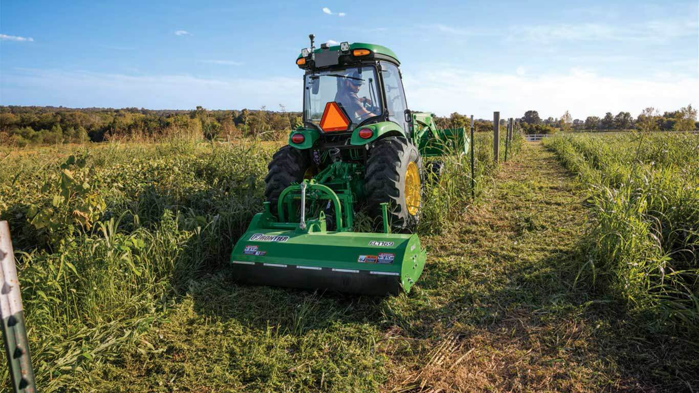 Green tractor with mower cuts tall grass in a field under clear sky.