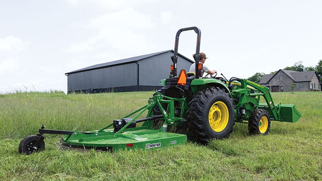 Tracteur vert avec faucheuse rotative fauchant l'herbe haute près d'une grande étable grise et d'une maison.