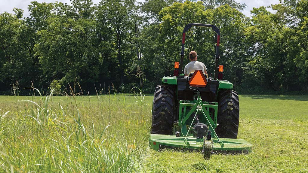 Un tracteur compact tire une faucheuse rotative verte à travers de l'herbe très haute dans un champ bordé d'arbres.