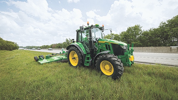 Tracteur vert tirant une tondeuse rotative sur le bord herbeux d'une route près d'une barrière en béton et sous un ciel nuageux