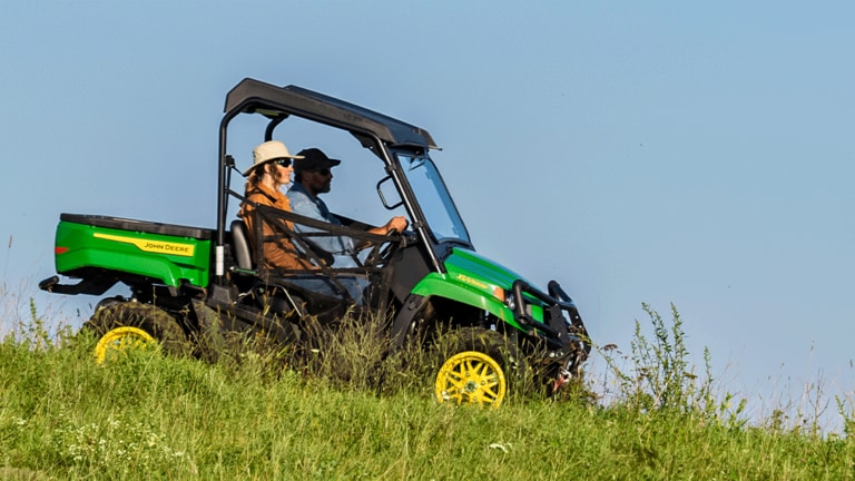Deux personnes conduisent un Gator vert en descendant une pente herbeuse sous un ciel dégagé.