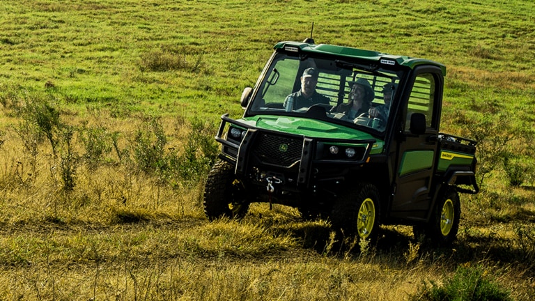 A green cabbed Gator carries three passengers through an open grassy field.