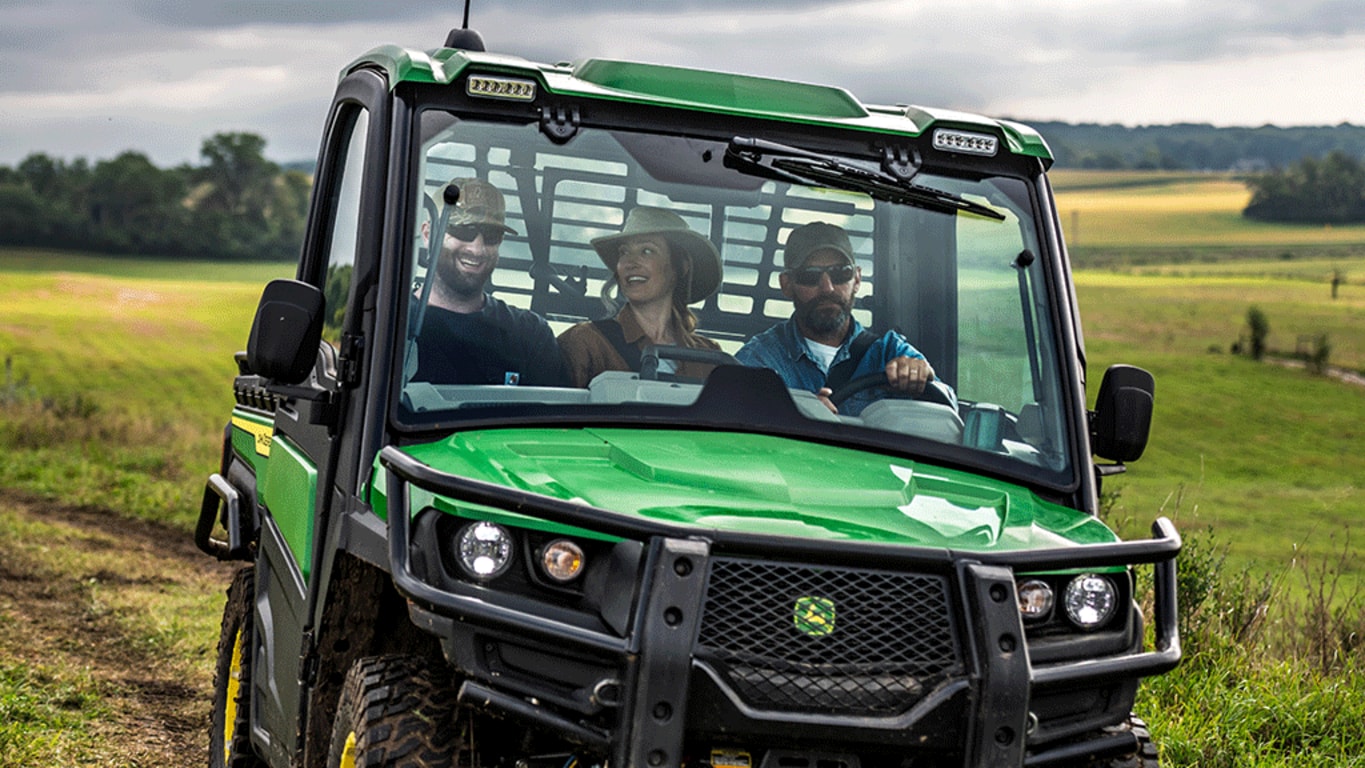 A close-up of a green cabbed Gator carrying three passengers through an open grassy field.