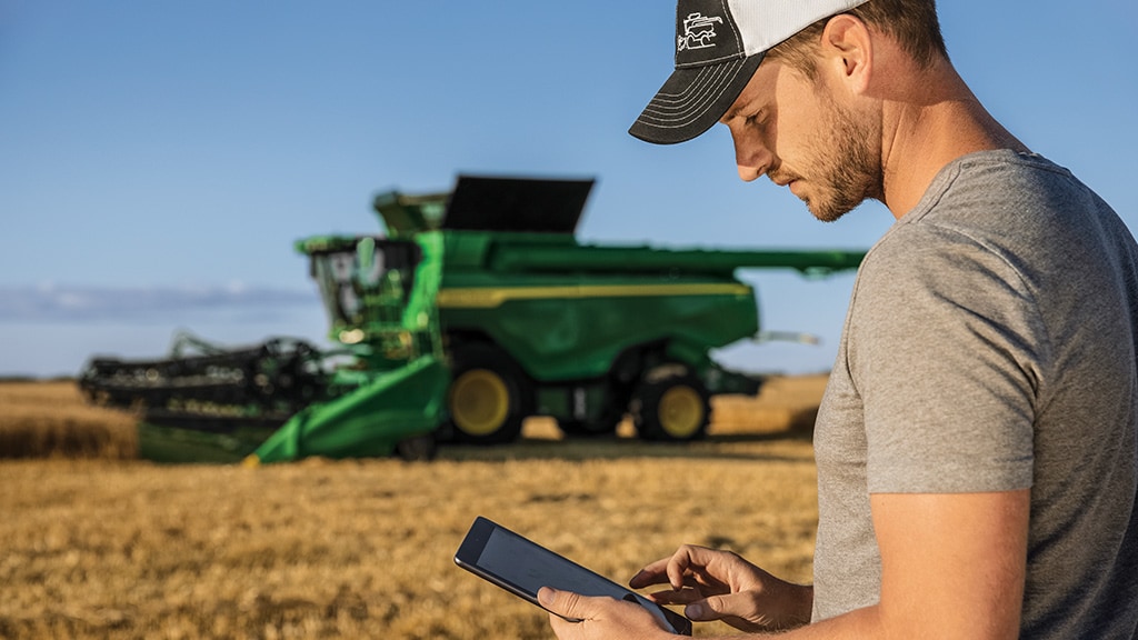 Photo d’un agriculteur debout devant une moissonneuse-batteuse S7 John Deere garée dans un champ de blé. L’agriculteur se sert de l’outil John Deere Connected Support sur son appareil mobile.