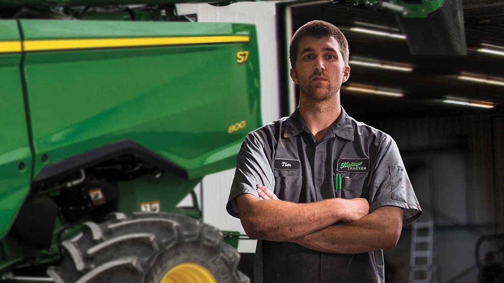 Photo of a John Deere technician standing in front of the rear of a John Deere Combine, in a shop, with his arms folded