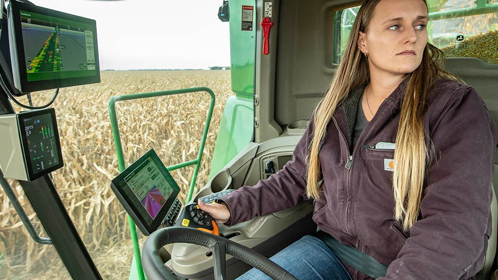 Photo à l’intérieur de la cabine d’une jeune agricultrice conduisant une moissonneuse-batteuse S7 John Deere dans un champ de maïs. Devant elle, vous pouvez voir un afficheur G5Plus, un module d’affichage principal sur montant de coin de nouvelle génération, et un afficheur supplémentaire G5Plus.