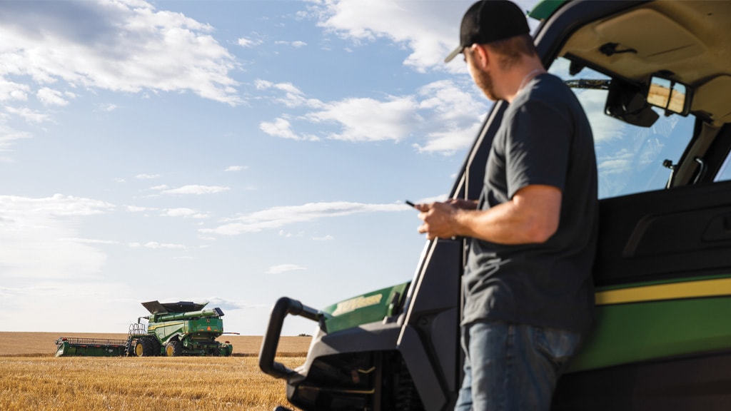 agriculteur appuyé contre un John Deere Gator, tout en vérifiant Le centre des opérations sur son appareil mobile. Vous pouvez voir un John Deere Combine en arrière-plan.