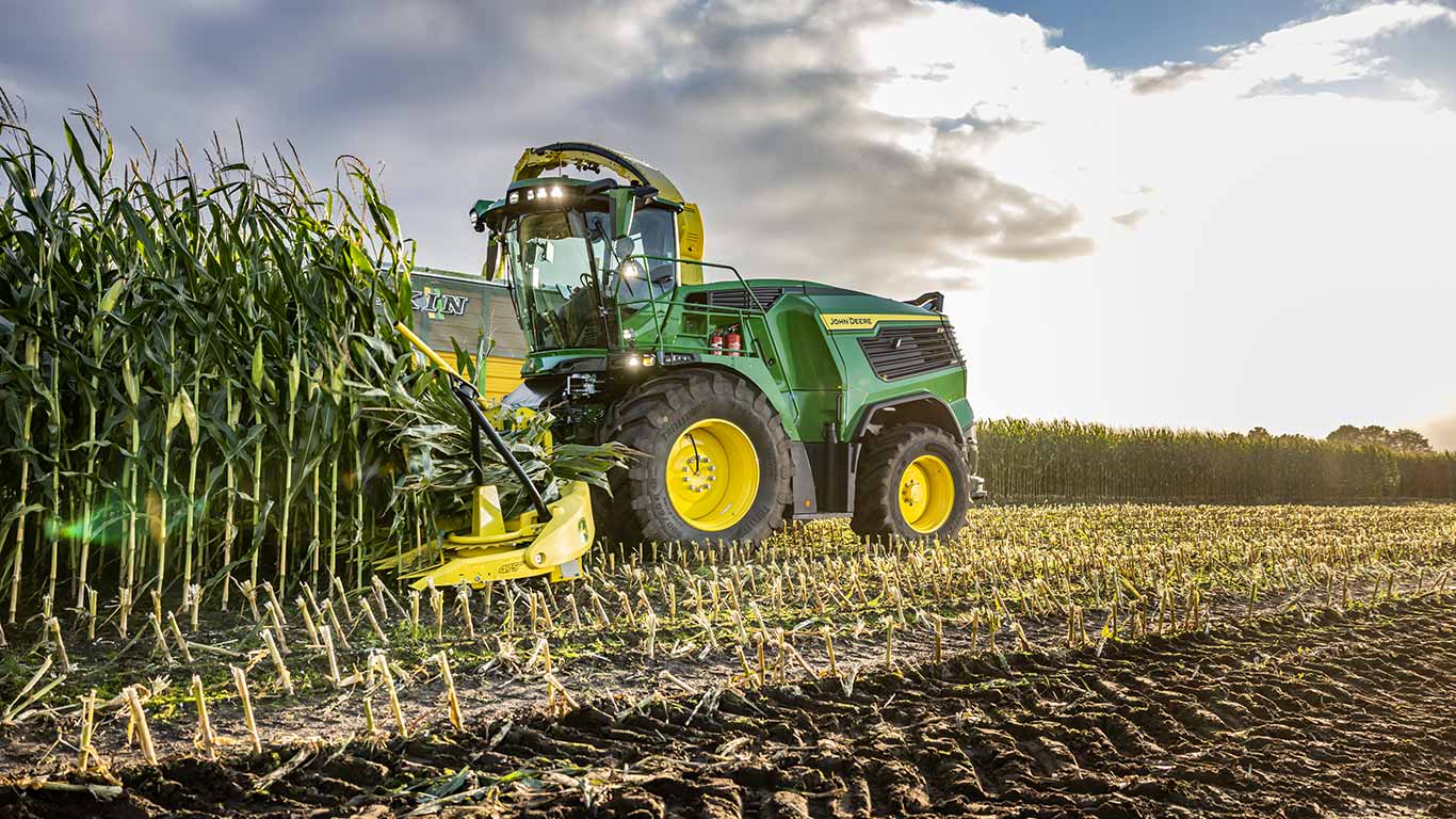Green John Deere tractor harvesting corn in a field under a partly cloudy sky.