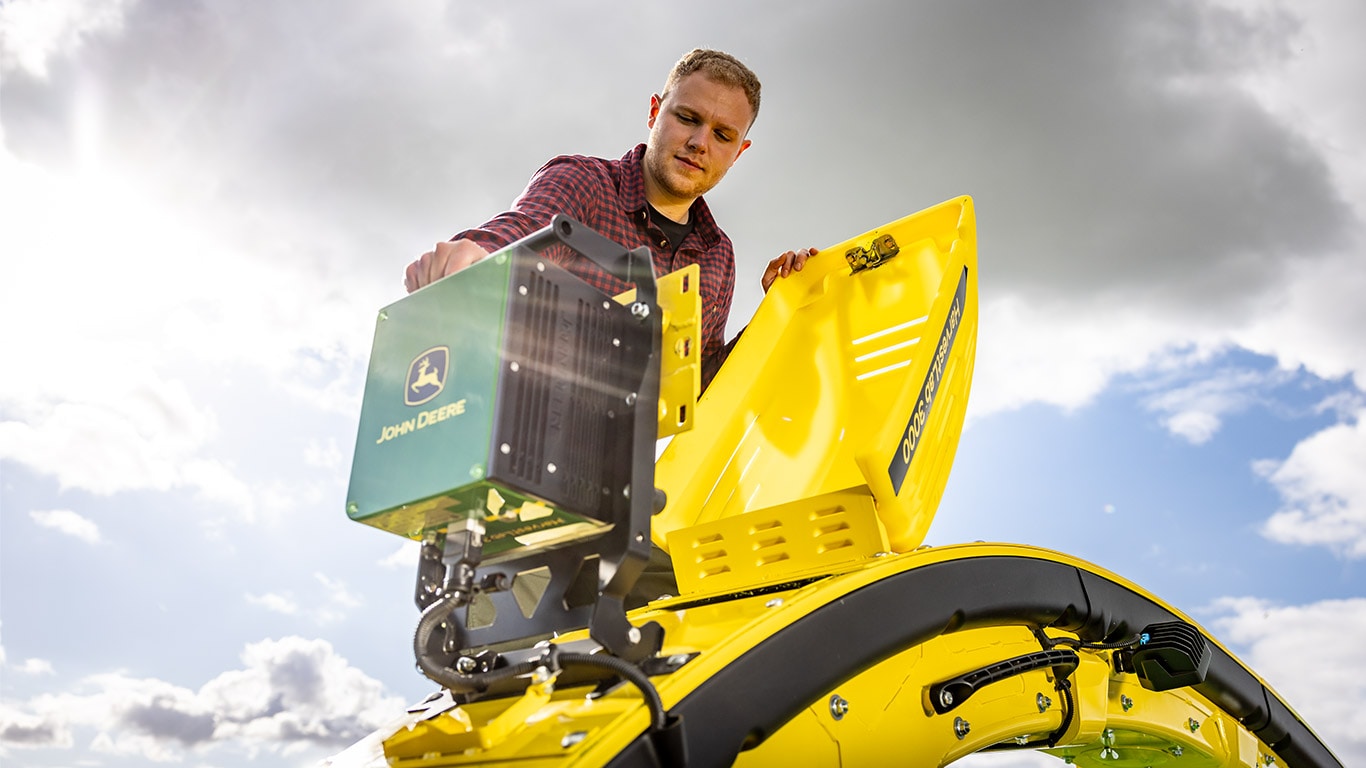 Person in red plaid shirt working on yellow John Deere machine under a cloudy sky.