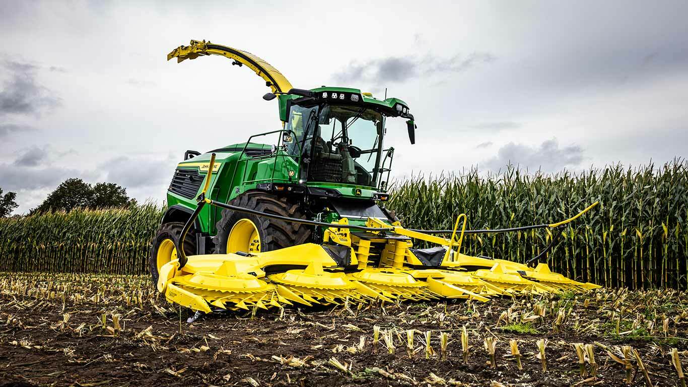 Green and yellow John Deere forage harvester in a cornfield under a cloudy sky.