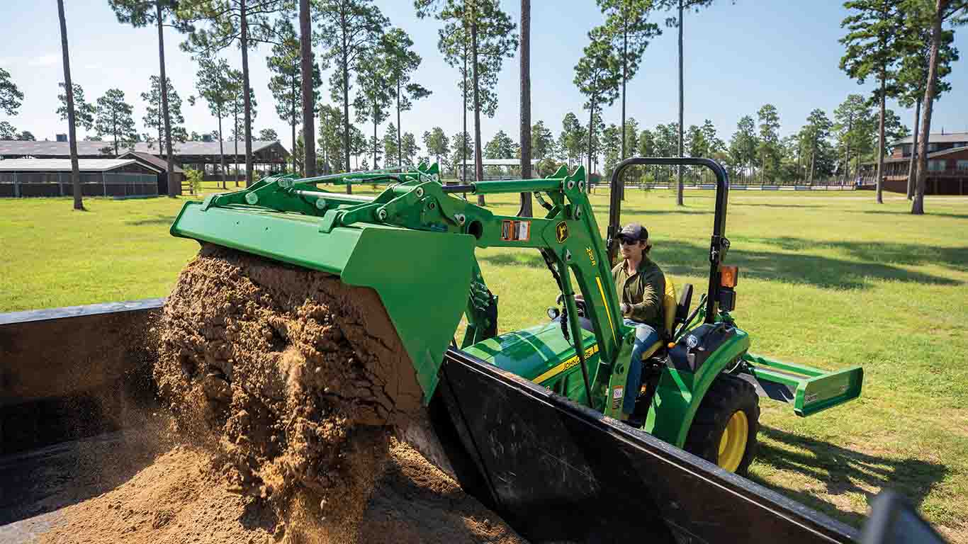 A green compact tractor with a bucket attachment in an open grassy area dumping soil into a black container.