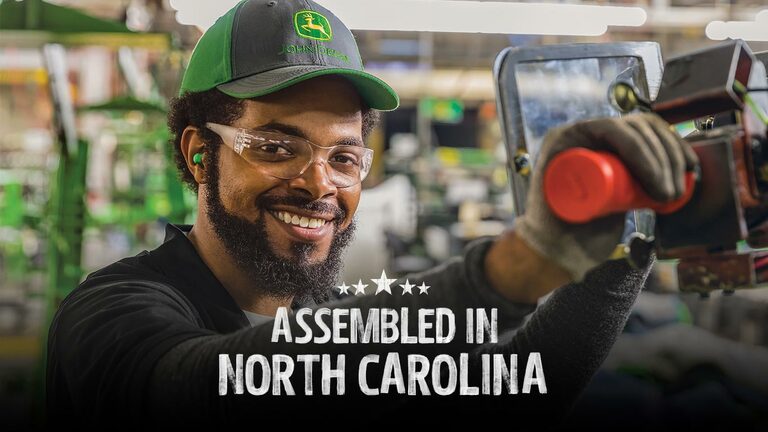 A smiling person safety glasses and a Deere hat assembling a piece of equipment at the factory in North Carolina.