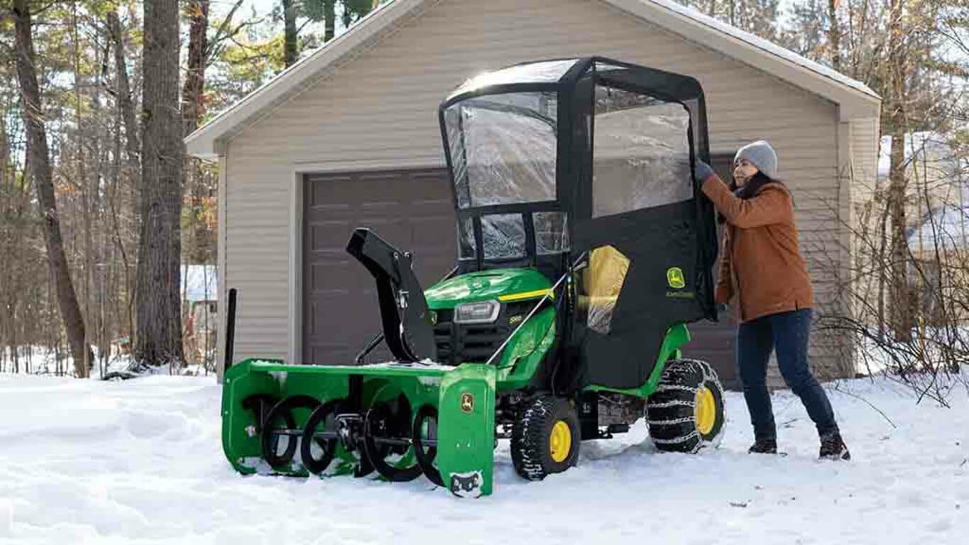 A green riding mower in snow with a snow blower and weather encloser attached to it.