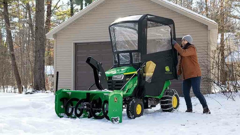 A green riding mower in snow with a snow blower and weather encloser attached to it.