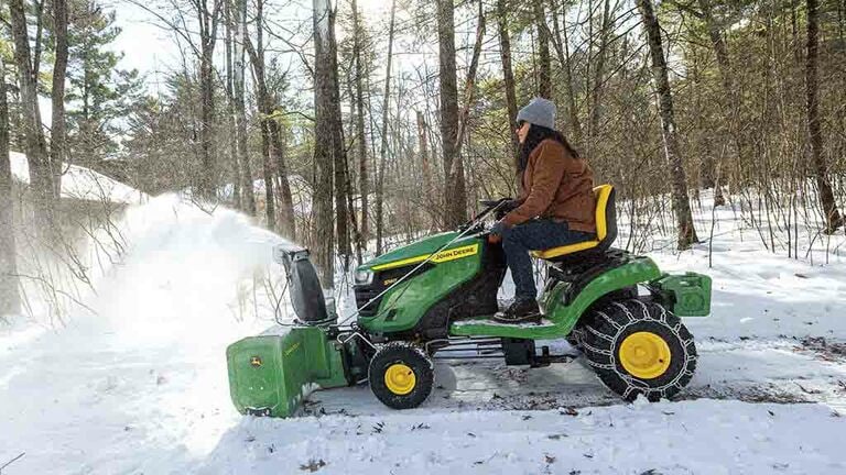 Snow being blown by a green riding mower equipped with a snow blower and snow chains.