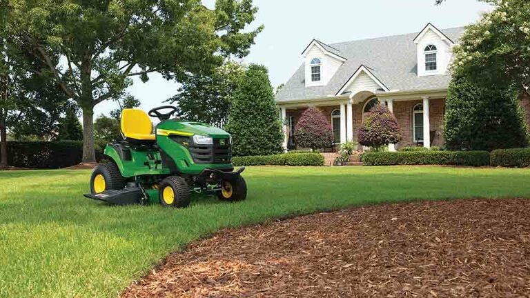 A rounded patch of mulch with a green riding mower next to it sitting on a grassy lawn.
