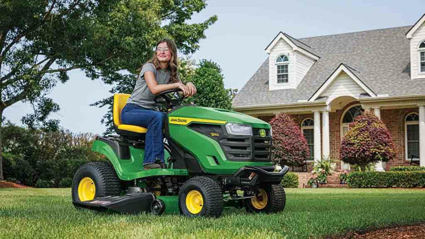 A smiling person resting on a green riding mower on a well-kept lawn.
