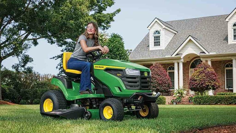 A smiling person resting on a green riding mower on a well-kept lawn.