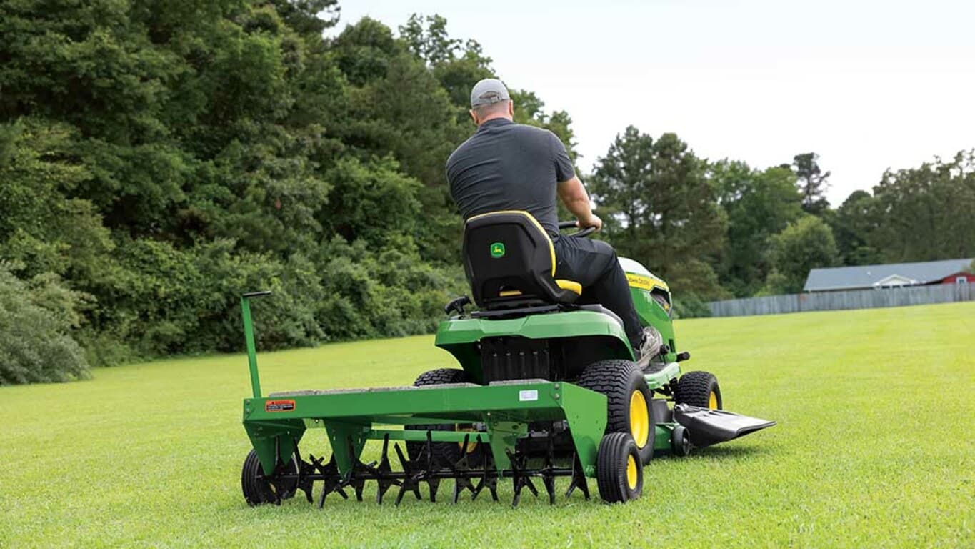 Rear view of a green lawn aerator being pulled by a green riding mower through an open grassy field.