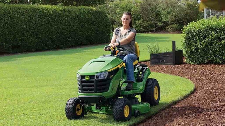 A person mowing the edge of a lawn by a patch of mulch on a green riding mower.