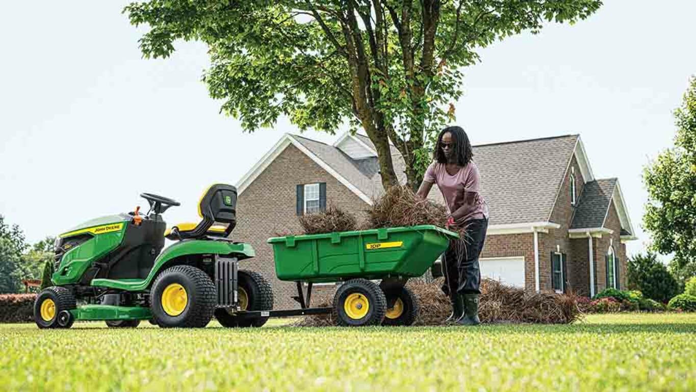 A person loading a green cart attached to the back of a green riding mower.