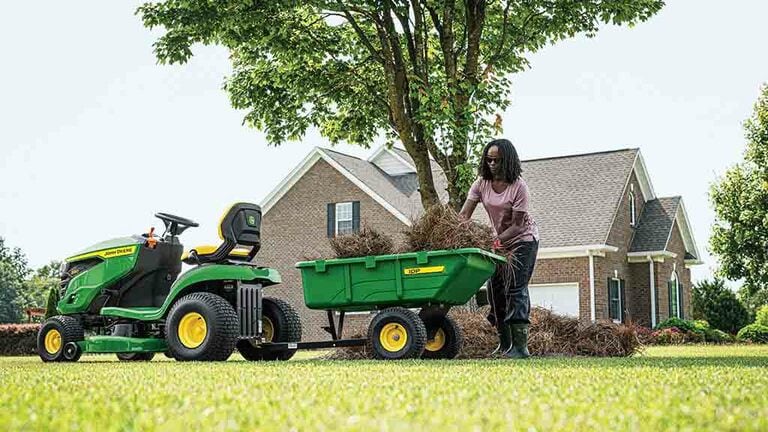 A person loading a green cart attached to the back of a green riding mower.