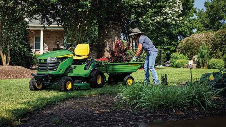 Plantes manipulées dans un chariot vert attaché à l'arrière d'une tondeuse autoportée verte.
